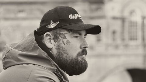 Cal in a cap, leaning on a railing by a stone bridge and looking out over the river