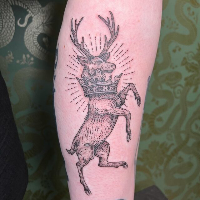 Etched tattoo of a rearing stag wearing a crown around its neck, with radiating lines, on a leg