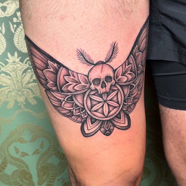 Moth with a skull on its body over a mandala of petals, tattooed across a thigh