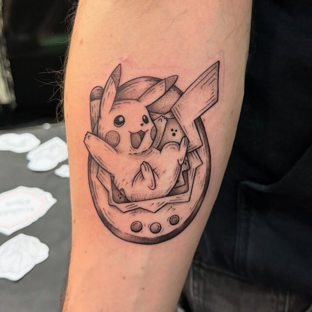 Pikachu bursting out of a lucky horseshoe in black and grey, tattooed on a forearm