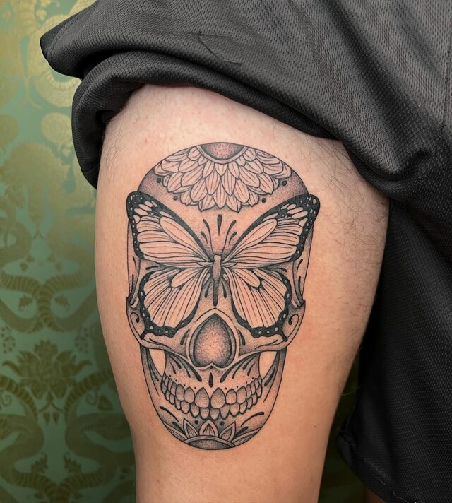 Skull with a butterfly spread across its eyes and a flower crown, tattooed on a thigh