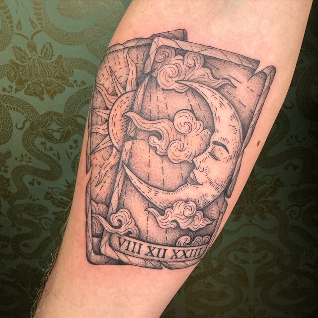 Tattoo of two tarot cards showing a sun and a crescent moon with a face among clouds, numbered in Roman numerals, on a forearm