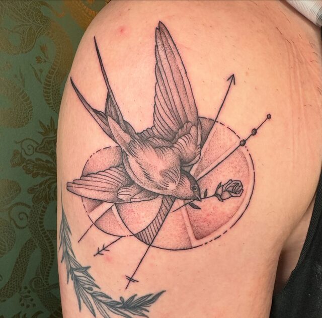 Swallow in flight carrying a rose, crossed by arrows over a dotwork circle, tattooed on a shoulder